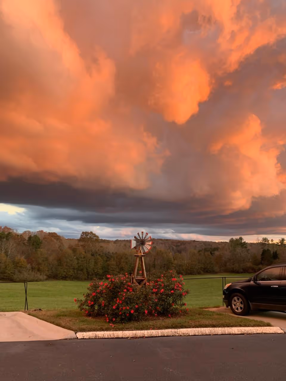 A scenic outdoor view at sunset with dramatic orange and dark clouds in the sky. In the foreground, there is a small decorative windmill surrounded by red flowering bushes, a grassy area, and a parked black SUV on the right side. Trees and a forested area are visible in the background.