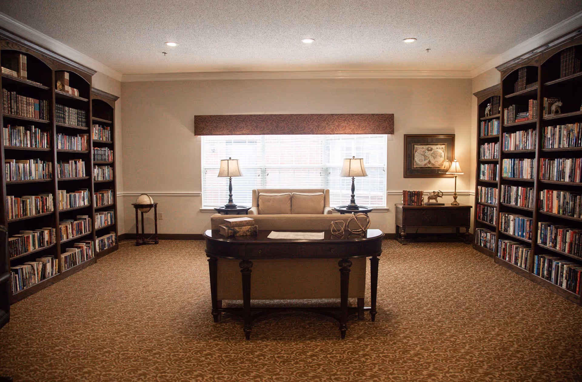 A cozy library room with tall wooden bookshelves filled with books on both sides. In the center, there is a beige sofa facing a dark wooden table with decorative items on it. Two table lamps are placed on side tables next to the sofa, and a large window with blinds is behind the sofa, allowing natural light to enter the room. The carpet has a patterned design, and the walls are painted a light color with crown molding.
