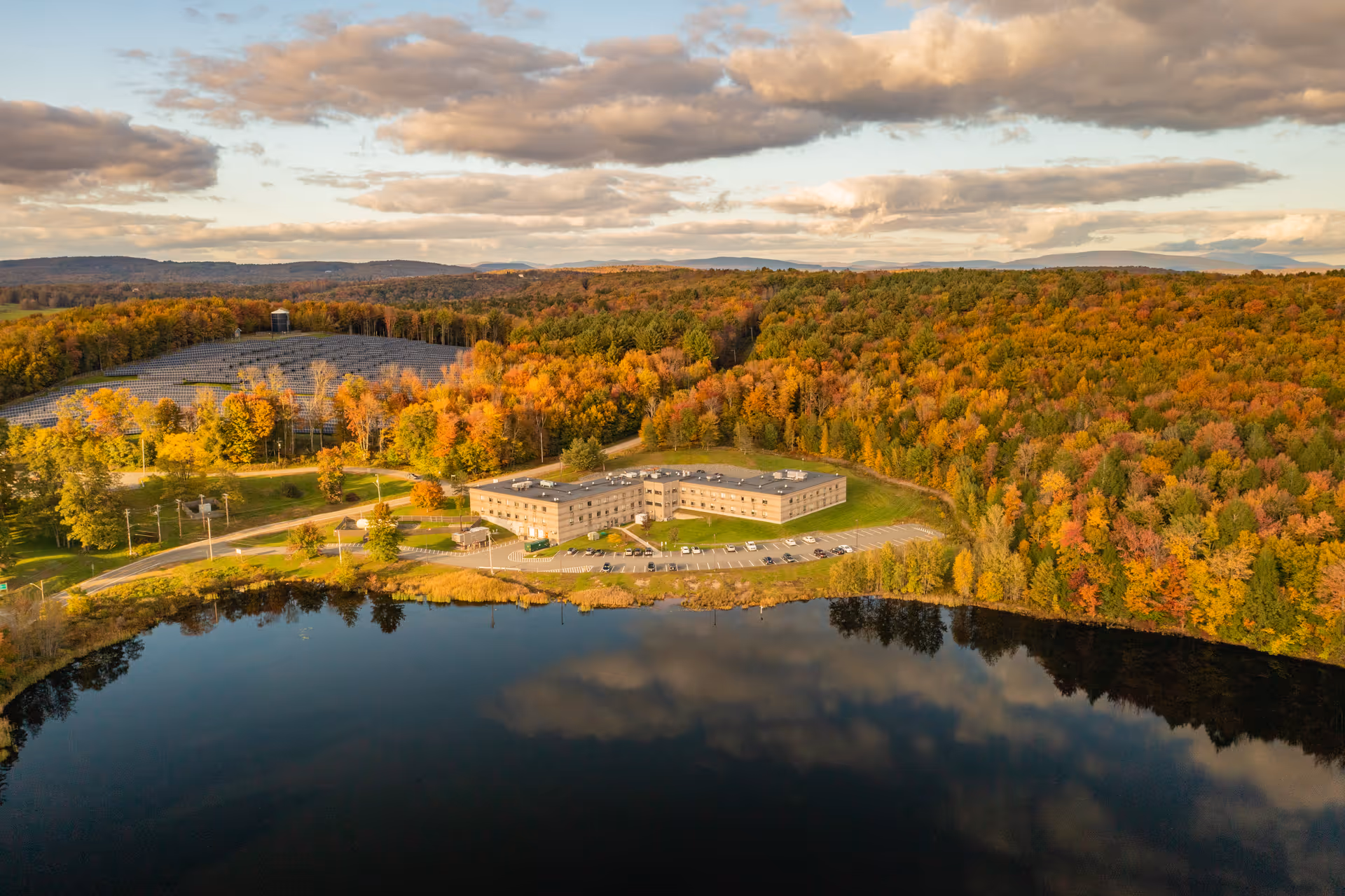 Aerial view of Care Center at Sunset Lake surrounded by dense forest with autumn foliage. The building is situated next to a calm lake reflecting the sky and clouds. There is a parking lot with cars in front of the facility and a solar panel installation visible on the left side among the trees.