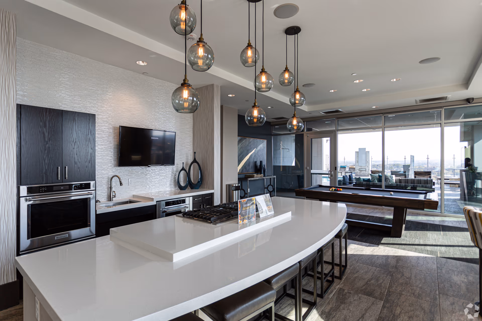 Modern kitchen area with a large white curved island countertop featuring a built-in gas stove and several stools underneath. Above the island, there are multiple hanging pendant lights with round glass bulbs. In the background, there is a wall-mounted flat-screen TV, dark wood cabinetry, a sink, and a dishwasher. To the right, a pool table is visible near large floor-to-ceiling windows that open to an outdoor seating area with a city view.
