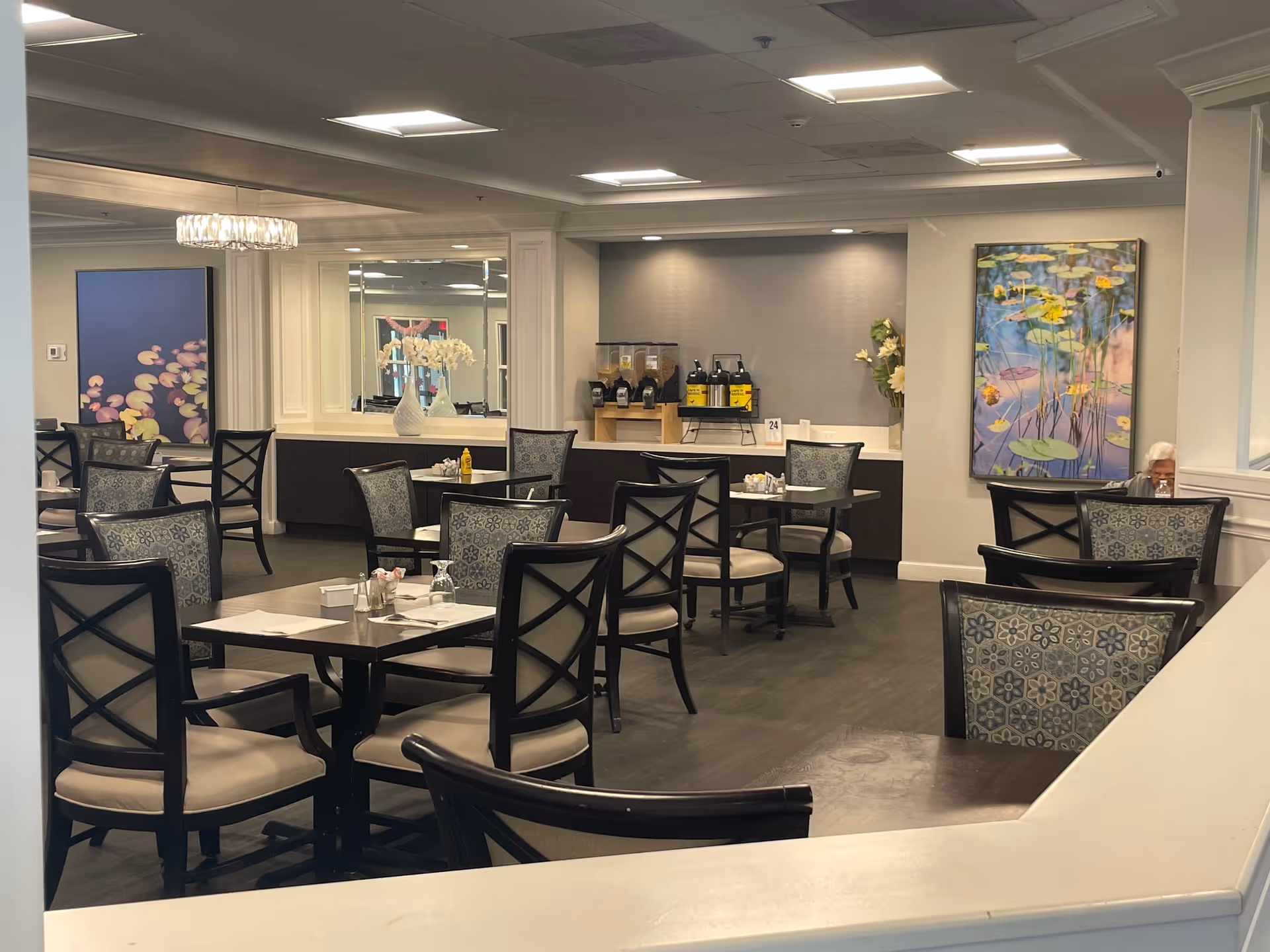 A dining room in a senior living facility with multiple tables and chairs arranged neatly. The room features decorative wall art, a large mirror, and a beverage station with coffee dispensers and condiments. There is one elderly person seated at a table near the window.