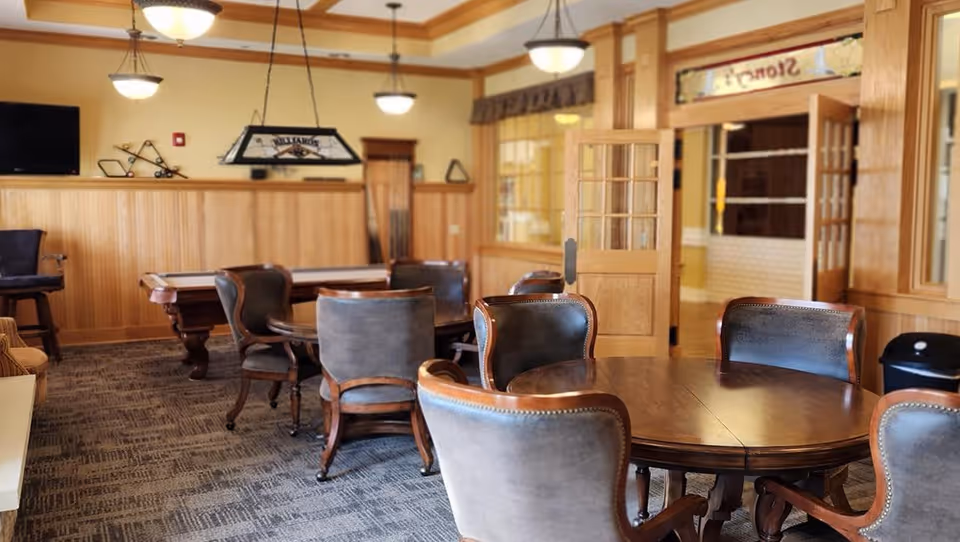 A cozy interior room with round wooden tables surrounded by cushioned chairs. The room features wood paneling on the walls, carpeted flooring, and ceiling lights. In the background, there is a pool table and a wall-mounted TV. A glass-paned wooden door is open, revealing another room with a sign above the door that reads 'Stoney'.