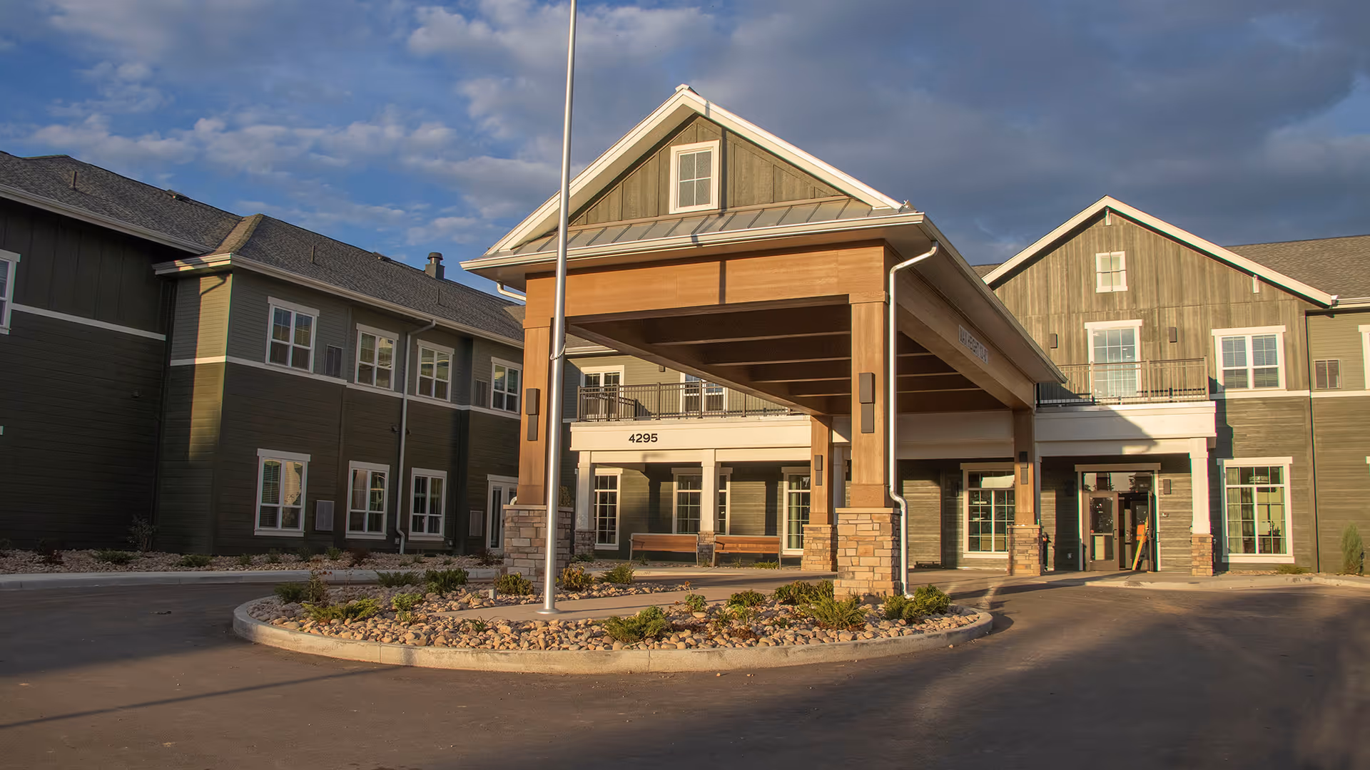 Exterior view of a senior living facility named Stoneridge at Centerra, showing a large covered entrance with wooden beams and stone pillars, surrounded by a paved driveway and landscaped area with small plants and rocks under a partly cloudy sky.