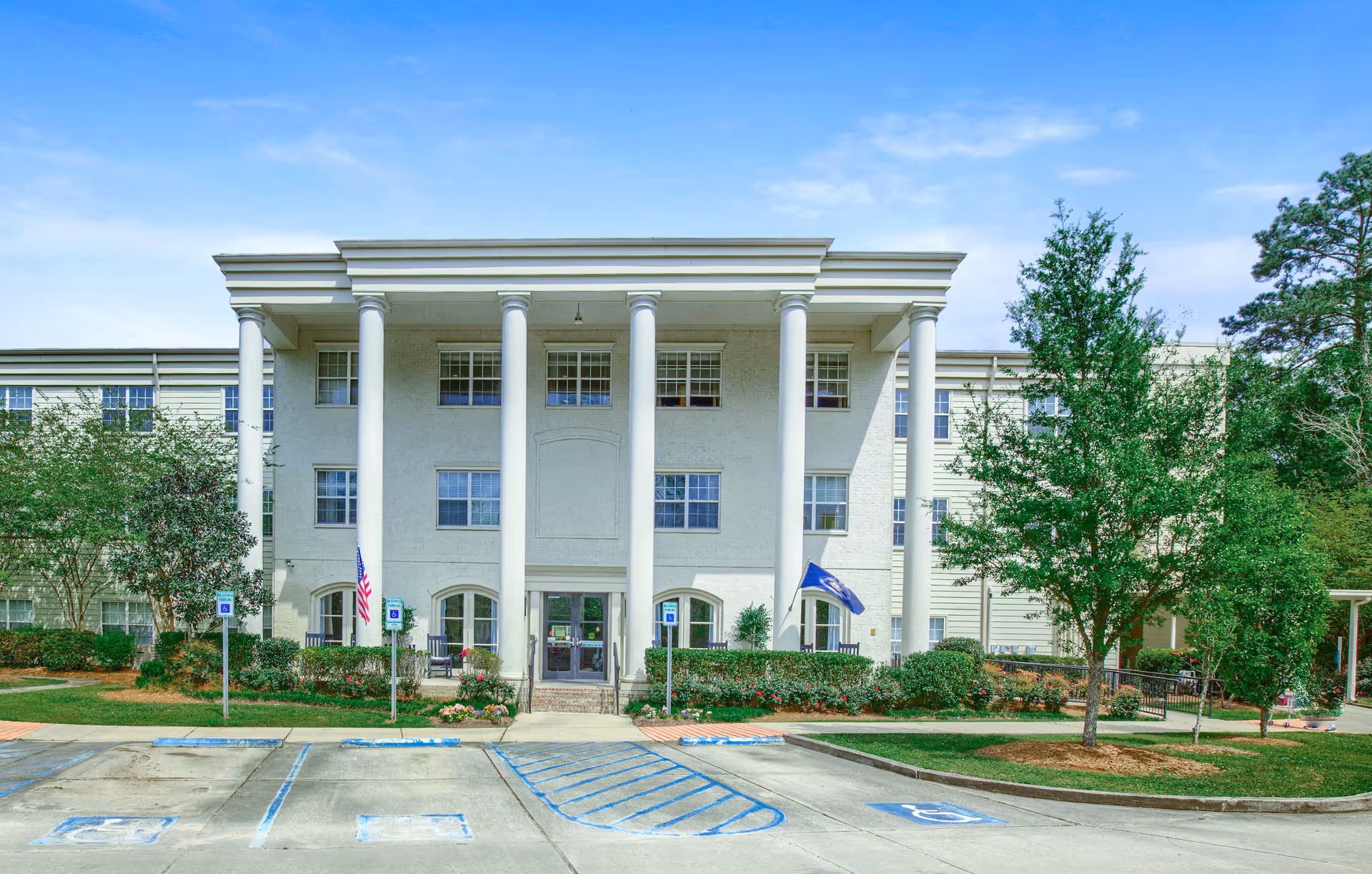 Front exterior view of a three-story senior living facility named The Trace, featuring large white columns, multiple windows, landscaped bushes and trees, and a parking area with handicap spaces in front.
