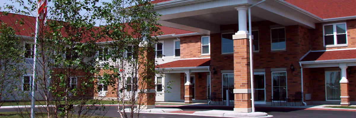 Front entrance of a two-story brick senior living building with a covered drop-off canopy and small trees in front.