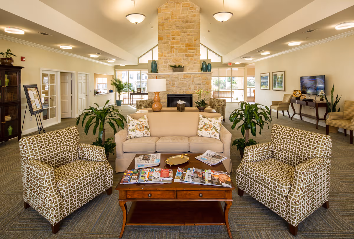 A spacious and well-lit living room area with a beige sofa and two patterned armchairs arranged around a wooden coffee table covered with magazines. Behind the sofa is a stone fireplace with decorative vases on the mantel. The room has large windows letting in natural light, several potted plants, framed pictures on the walls, and additional seating along the sides.