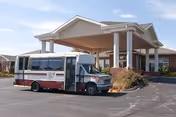 A small shuttle bus parked under the covered porte-cochère at the entrance of a single-story brick senior living building.