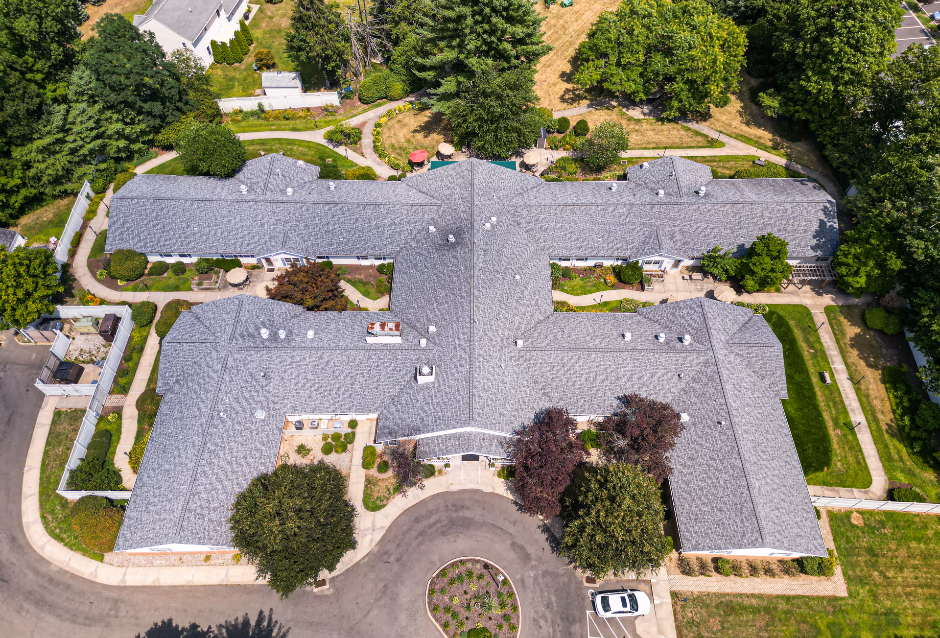 Aerial view of Arden Courts - ProMedica Memory Care Community in Farmington showing a large, single-story building with a gray roof surrounded by landscaped gardens, trees, pathways, and a circular driveway with a parked white car.