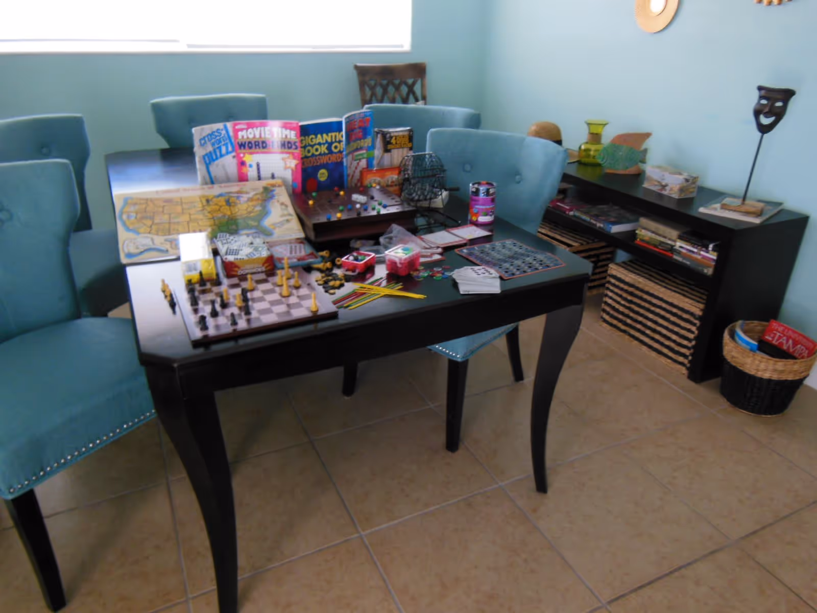 Table covered with board games, a chess set and puzzles surrounded by teal chairs in a light-blue activity room.