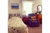 A cozy bedroom with a single bed covered in a floral bedspread, a wooden nightstand with a lamp and picture frames, a comfortable blue recliner chair with a decorative pillow, a wooden dresser with a mirror, and a window with a valance letting in natural light.