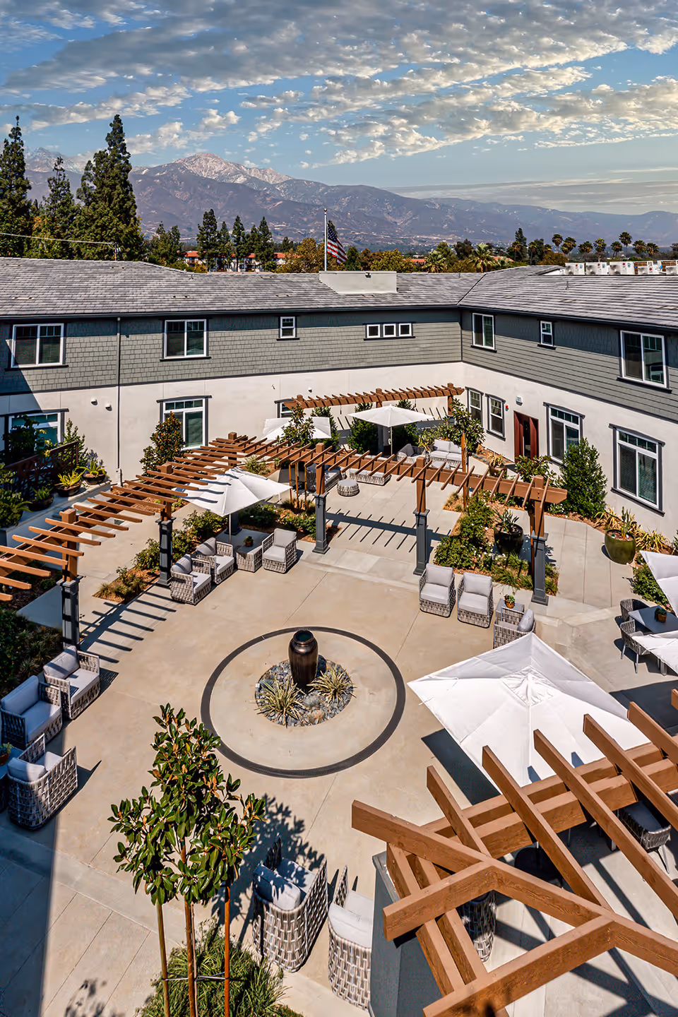 A spacious outdoor courtyard at Allara Senior Living featuring comfortable seating areas with cushioned chairs and umbrellas, wooden pergolas providing partial shade, a central decorative water feature, and surrounding greenery. The courtyard is enclosed by a two-story building with multiple windows, and mountains are visible in the background under a partly cloudy sky.
