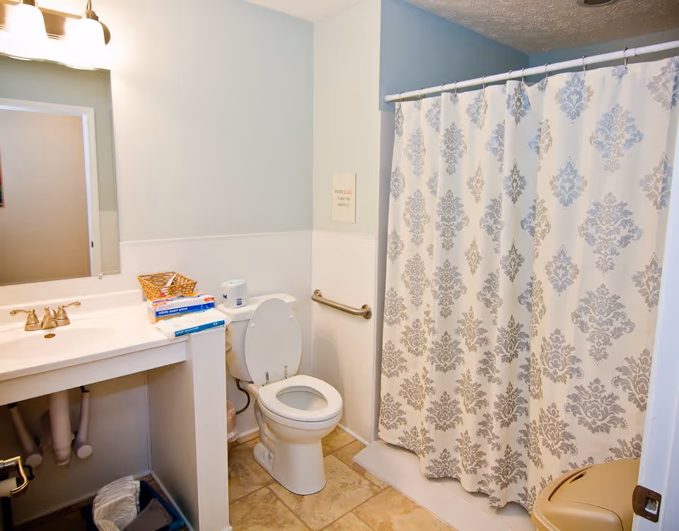 A clean bathroom with a white toilet, a sink with a countertop holding a basket and some boxes, a shower with a patterned curtain, a grab bar on the wall, and a trash can on the floor.