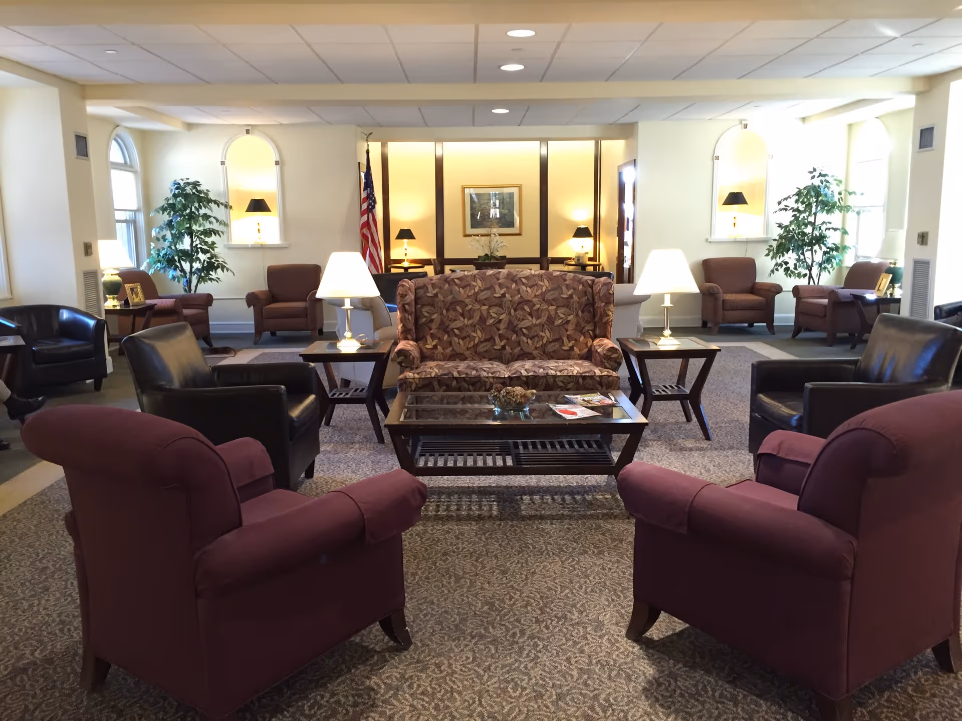 A spacious senior living facility common area with multiple armchairs and a patterned loveseat arranged around a glass coffee table. The room is well-lit with table lamps and natural light from windows. There are decorative plants, framed artwork, and an American flag in the background.