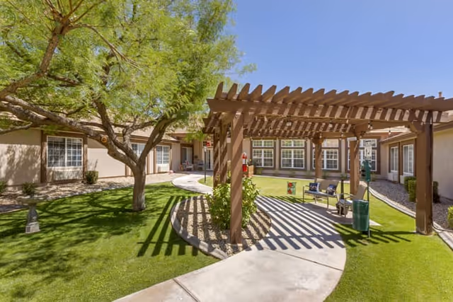 Sunlit courtyard with a wooden pergola, seating area, curved walkway, green lawn and surrounding single-story building.
