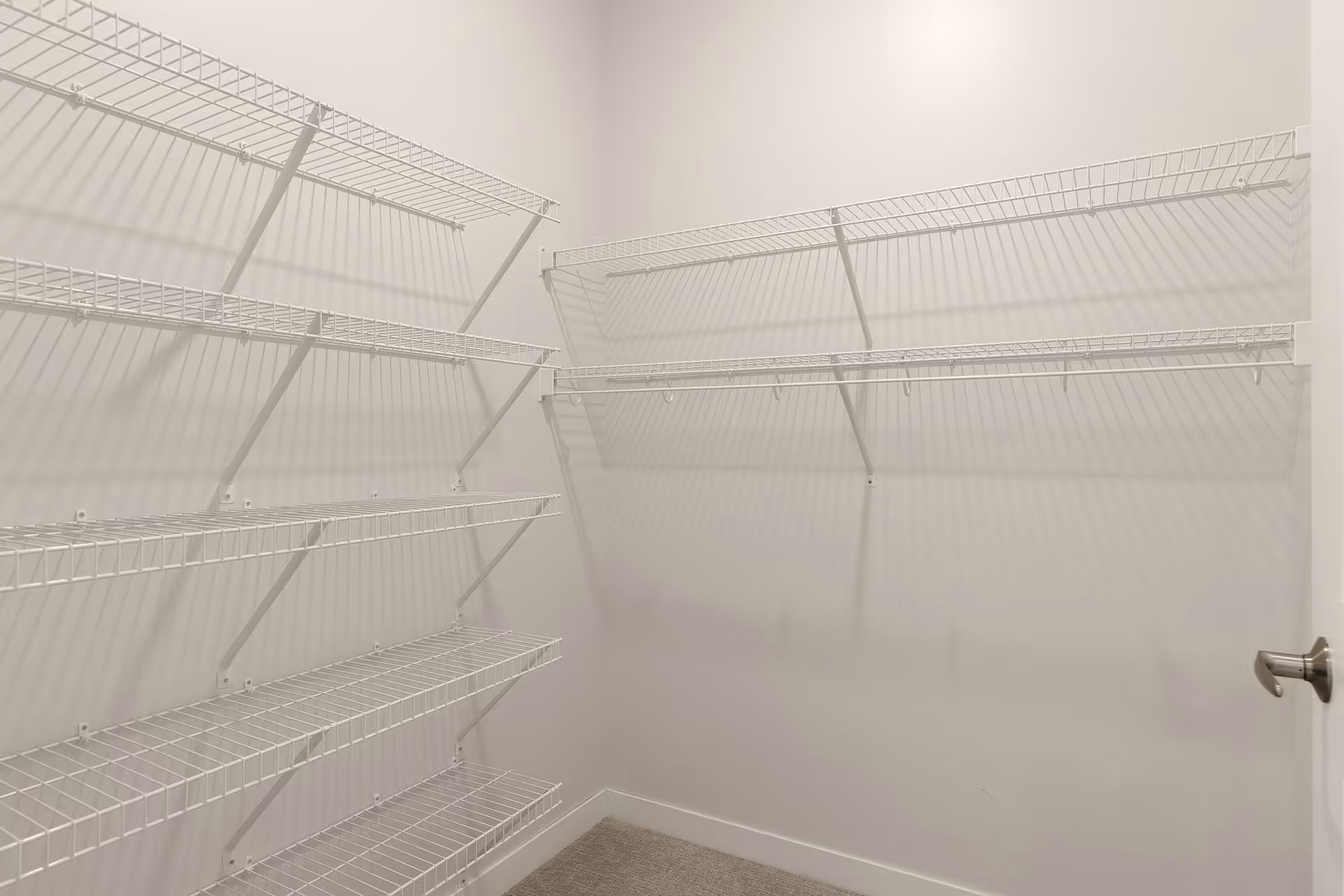 Empty walk-in closet with multiple white wire shelves mounted on two adjacent walls and a beige carpeted floor.