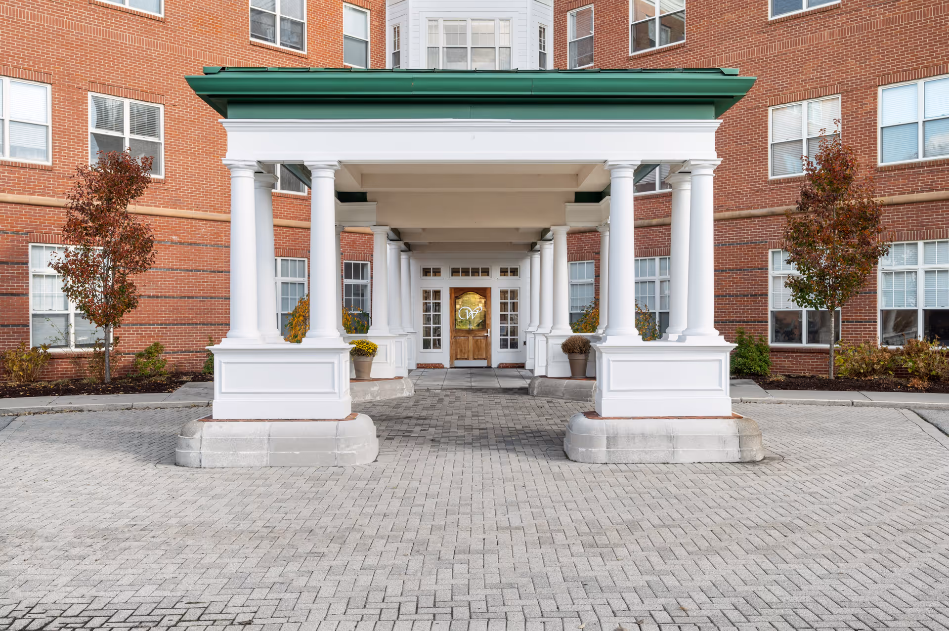 Entrance of Wingate Residences on Blackstone Boulevard showing a covered driveway with white columns and a green roof. The building is made of red brick with multiple windows, and there are small trees and plants on either side of the entrance.