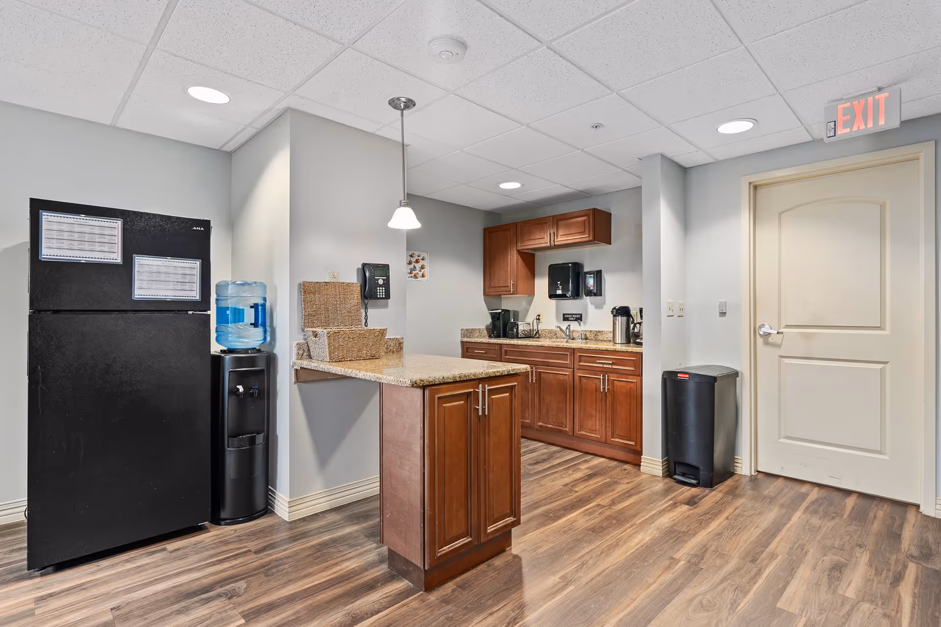 Interior view of a small kitchen area with wooden cabinets, a black refrigerator, a water dispenser, a countertop island, a coffee maker, a sink, and a trash bin near a closed door with an exit sign above it.
