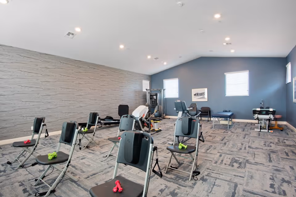 A spacious fitness room with several exercise chairs equipped with resistance bands and small dumbbells. The room has a patterned carpet, light gray textured wall on the left, and blue walls with windows on the right. There is additional exercise equipment and a treatment table near the back wall.