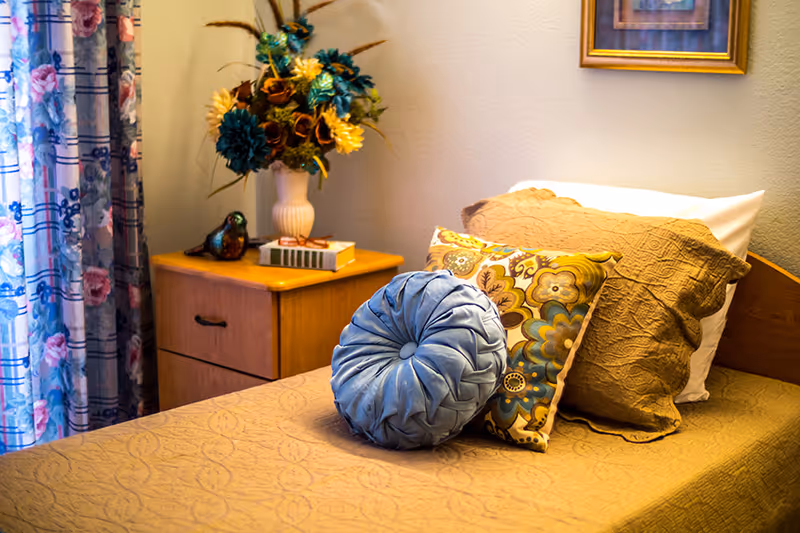 A neatly made single bed with a beige quilted bedspread and three decorative pillows in blue and floral patterns. Next to the bed is a wooden nightstand with a vase of colorful artificial flowers, a small decorative bird figurine, and a couple of books. Floral patterned curtains hang beside the nightstand, and a framed picture is partially visible on the wall above the bed.