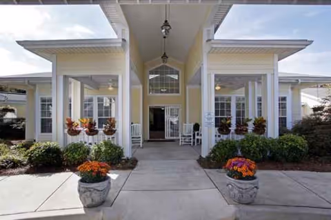 Front entrance of a light-yellow senior living facility with columns, hanging lights, and potted flowers.