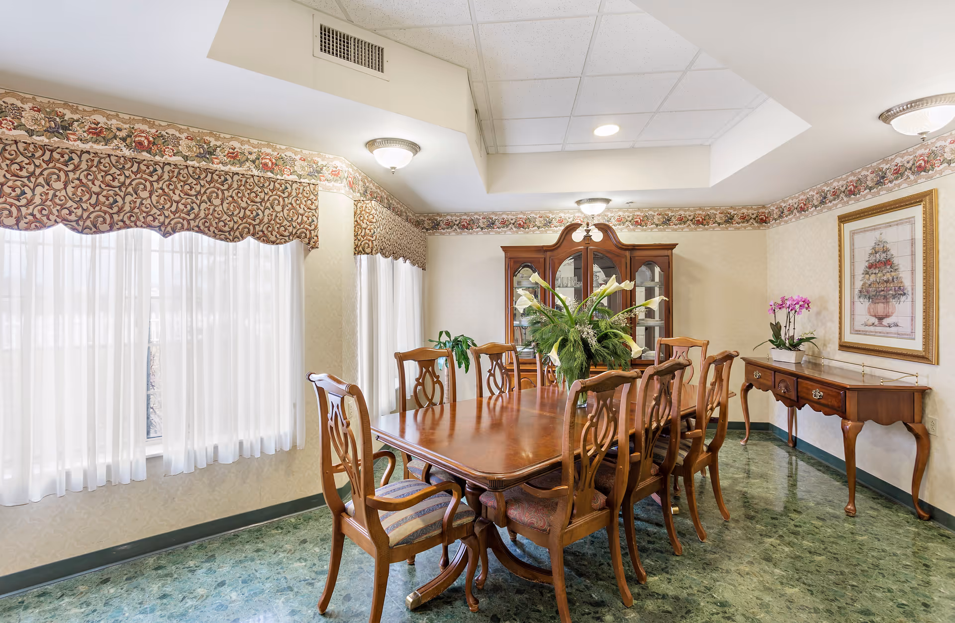 A formal dining room with a large wooden dining table surrounded by eight matching chairs. The room features floral valances over sheer white curtains on the windows, a wooden china cabinet with glass doors, a side table with a potted orchid, and framed artwork on the wall. The floor has a green marble-like pattern, and the ceiling has recessed lighting and a decorative light fixture.