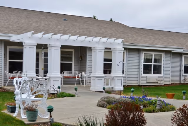 Outdoor patio area of a senior living facility named Palouse Hills featuring white pergola columns, white metal chairs, a butterfly-shaped bench, potted plants, and a garden with flowers and shrubs in front of a single-story building with gray siding and multiple windows.