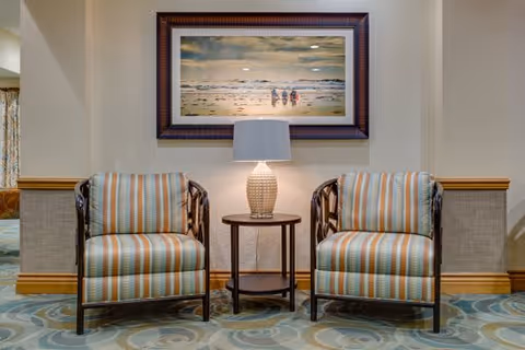 Two striped armchairs with wooden frames are placed on either side of a small round wooden table with a textured lamp on it. Above the table hangs a framed painting depicting a beach scene with three people walking along the shore. The room has patterned carpet and light-colored walls with wooden trim.