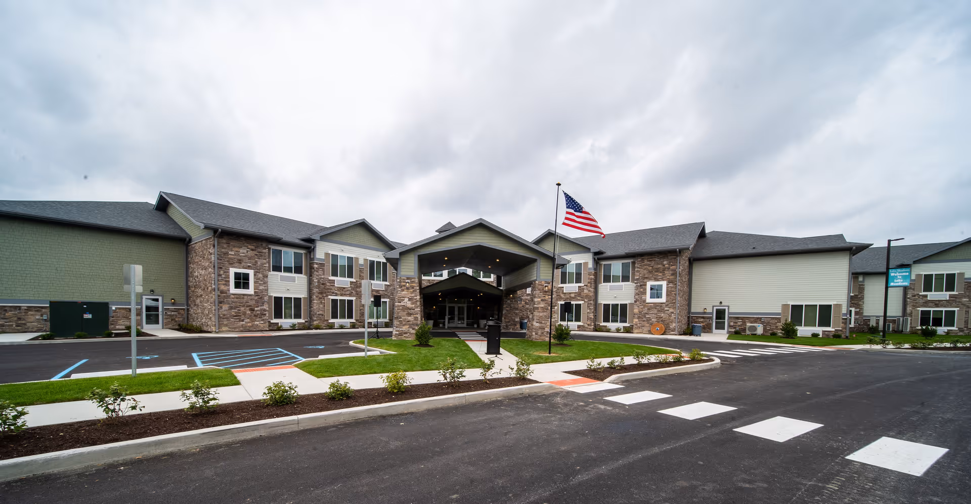 Front exterior view of Lake Meadows Assisted Living facility showing a two-story building with a covered entrance, an American flag on a flagpole, parking spaces including handicapped spots, landscaped areas with small bushes, and a cloudy sky overhead.