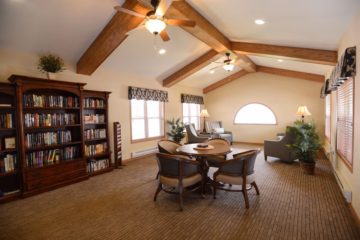 A spacious living room with vaulted ceilings featuring exposed wooden beams and ceiling fans. The room includes a large wooden bookshelf filled with books, a round table with four chairs in the center, and several armchairs arranged near windows with patterned valances. There are also potted plants and table lamps providing a cozy atmosphere.