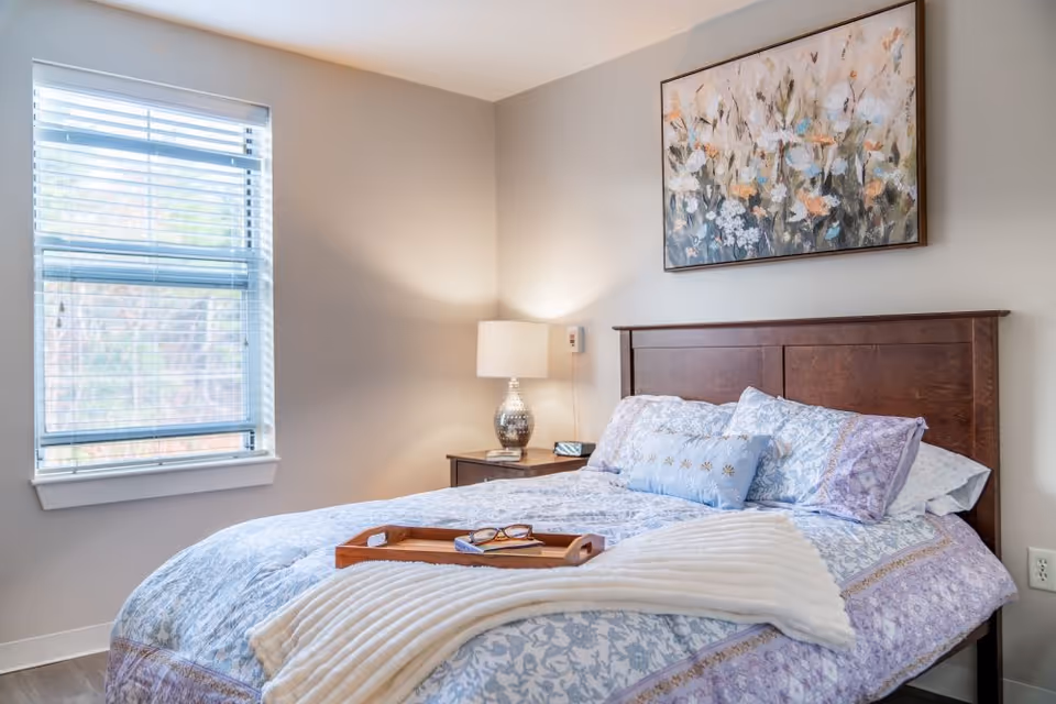 A cozy bedroom with a wooden headboard bed dressed in light blue and purple floral bedding, a white throw blanket, and decorative pillows. A wooden tray with glasses and a book rests on the bed. To the right of the bed is a nightstand with a silver lamp and a small clock. A large window with white blinds is on the left wall, and a floral painting hangs above the bed.