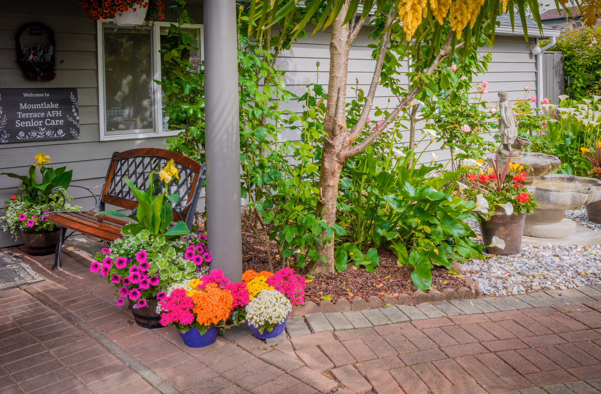 Entrance garden area of Mountlake Terrace Adult Family Home with a bench, colorful potted flowers, a small fountain, and a welcome sign on the building.