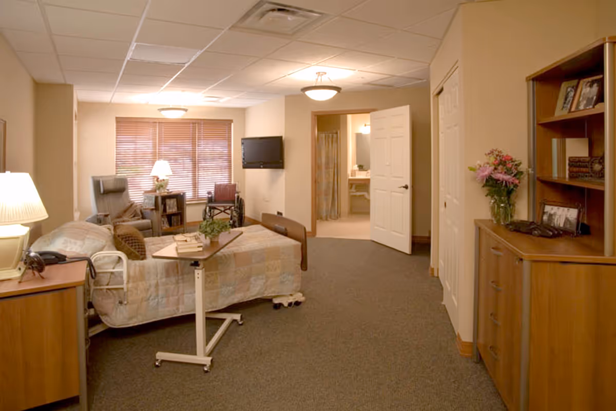 A cozy senior living bedroom with a hospital-style bed covered with a quilt, a small overbed table with books and a plant, a wheelchair, an armchair, a side table with a lamp, a wall-mounted TV, and a window with wooden blinds. The room has beige walls and carpeted floor, with a door open to a bathroom in the background. There is a wooden dresser with flowers and framed photos on the right side.