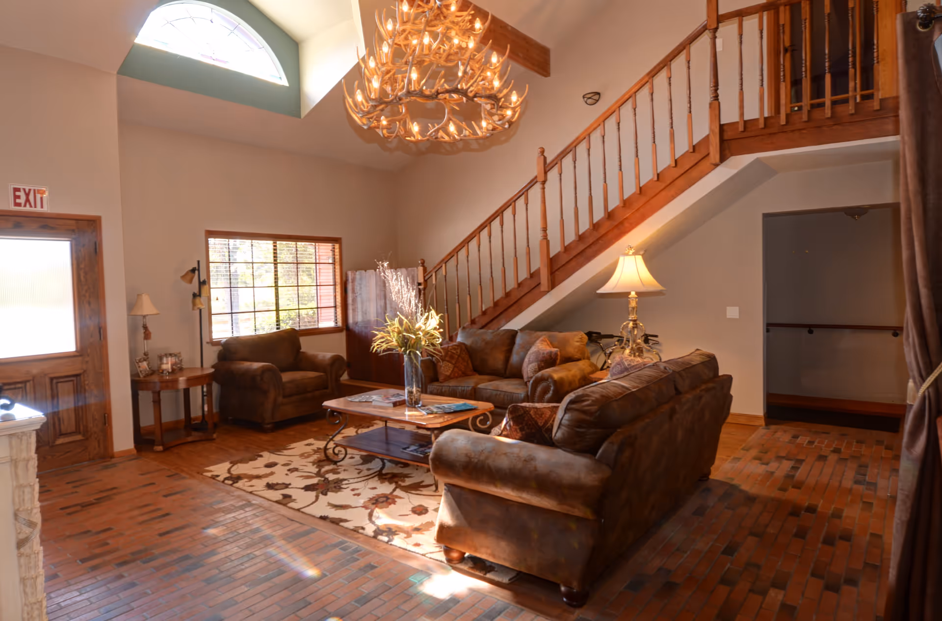 A cozy living room area with brown leather sofas and armchairs arranged around a wooden coffee table with a vase of flowers. The room features a brick floor, a large window, a wooden staircase with a railing, and a decorative chandelier hanging from a high ceiling. A table lamp is placed on a side table next to one of the sofas.