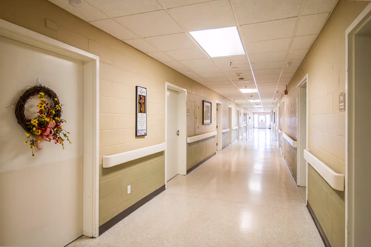 A long, well-lit hallway in a healthcare or senior living facility with beige and light green walls, white doors, handrails on both sides, and a decorative wreath hanging on one door.