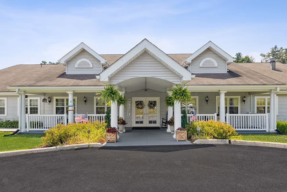 Front exterior view of a single-story building with a covered entrance supported by white columns, hanging green plants, and a welcome sign next to double glass doors. The building has gray siding, white trim, and a shingled roof with dormer windows. There are small American flags and flower pots near the entrance, and a paved driveway in front.