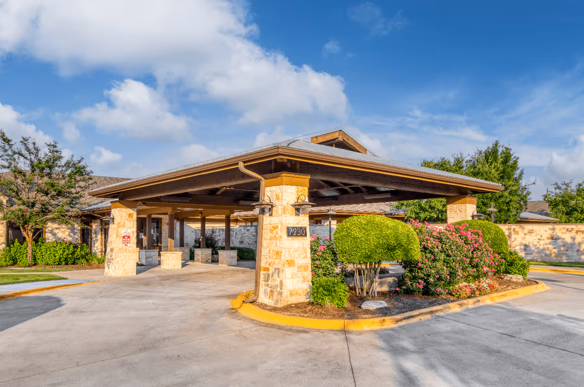 Entrance of The Waterford at Round Rock facility showing a covered drop-off area with stone pillars, neatly trimmed bushes, flowering plants, and a clear blue sky with some clouds.
