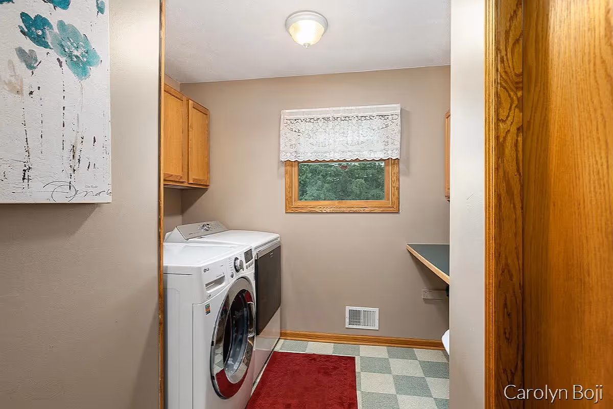Laundry room with a front-loading washing machine and dryer side by side, wooden cabinets above, a small window with a lace curtain, a red rug on the checkered floor, and beige walls.