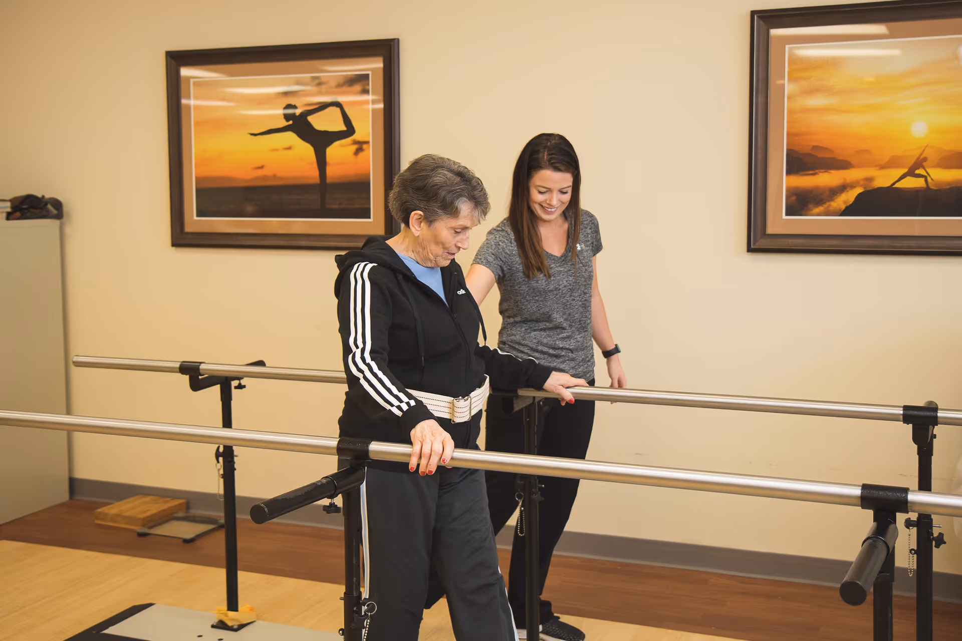 An elderly woman using parallel bars for physical therapy, assisted by a younger female therapist in a rehabilitation room with framed pictures of yoga poses on the wall.