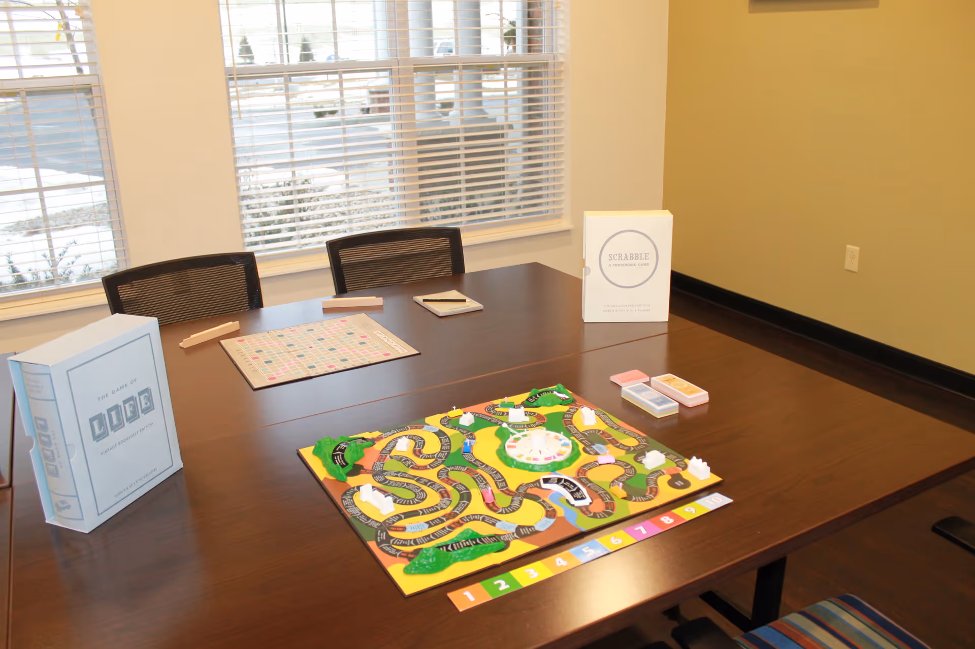 Large wooden table in an activity room set up with board games and chairs by a window.
