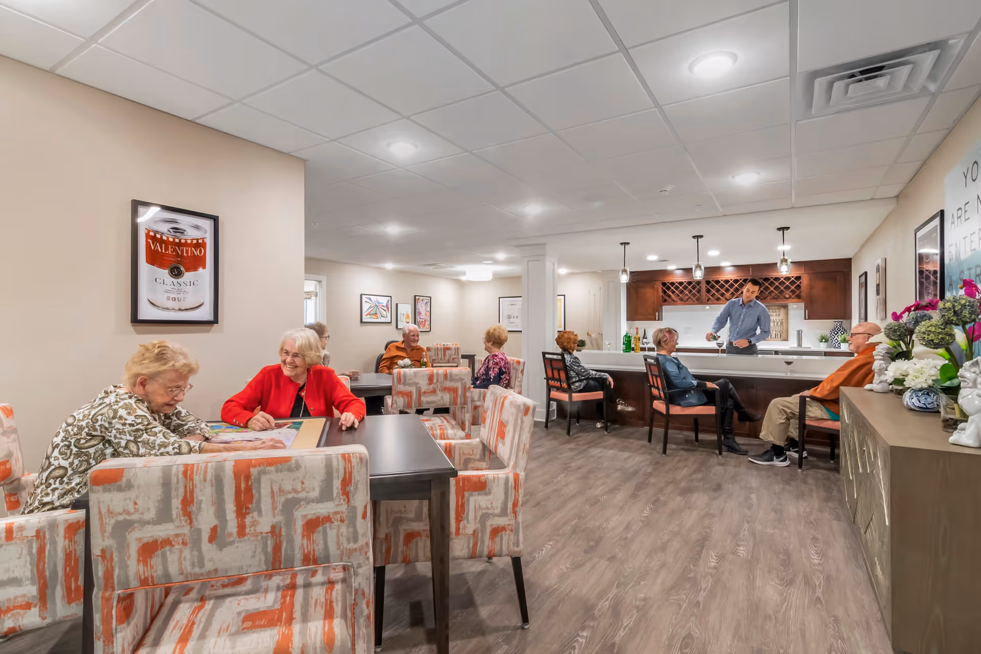 A senior living community common area with elderly residents sitting and socializing at tables and a bar counter. The room features patterned chairs, wood flooring, framed artwork on the walls, and a man behind the bar serving drinks. The atmosphere is warm and inviting with ceiling lights and decorative flowers on a side cabinet.