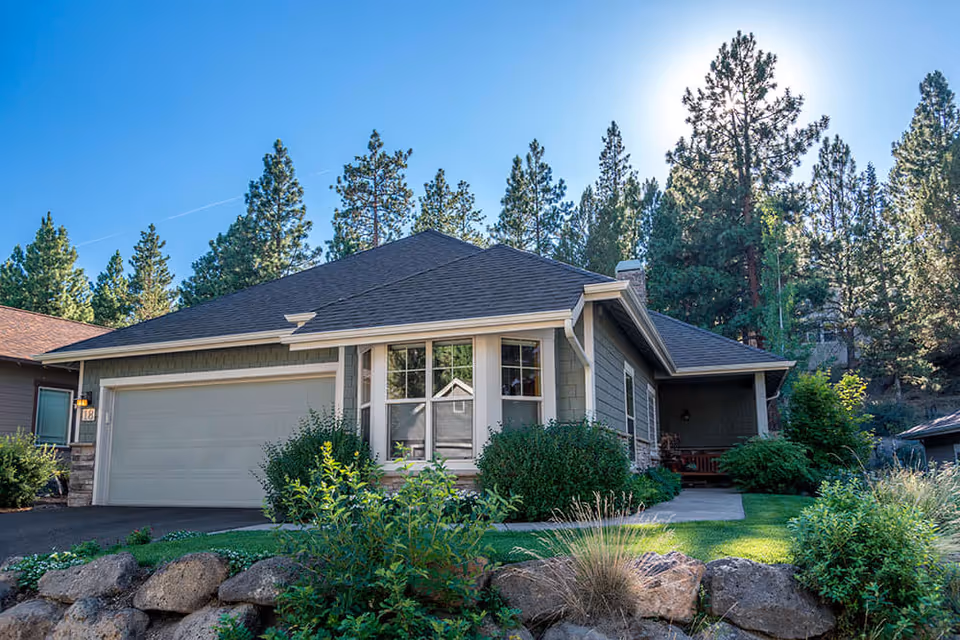Exterior view of a single-story residential building with a two-car garage, surrounded by greenery and tall pine trees under a clear blue sky with the sun shining.