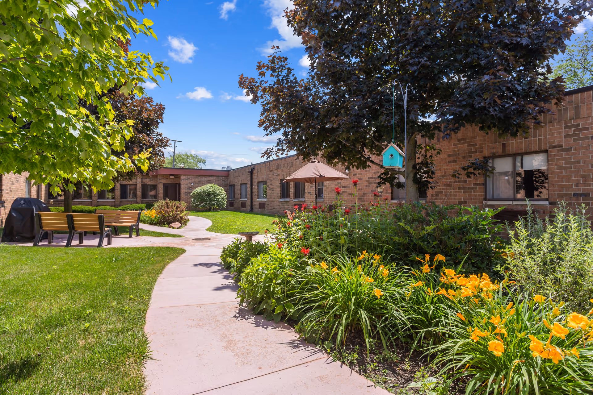 Outdoor garden area at Farmington Hills Inn Assisted Living with a paved walkway, green grass, colorful flowers, trees, benches, a covered grill, and a birdhouse hanging from a tree. The building with brick walls and windows surrounds the garden area under a blue sky with some clouds.