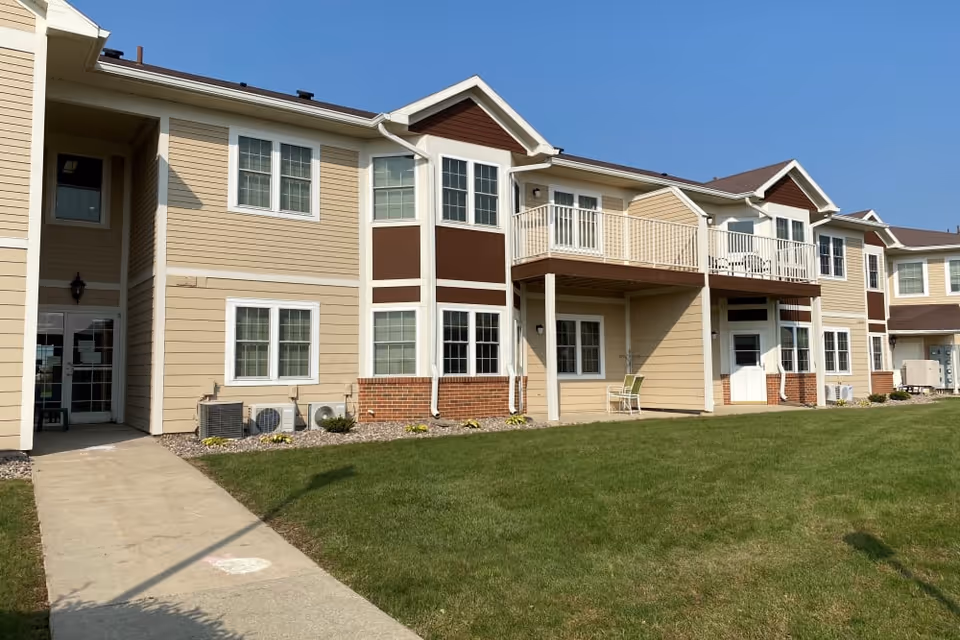 Exterior view of a two-story senior living facility building with beige siding, white trim, and brown accents. The building features multiple windows, a balcony, a patio area with chairs, and a concrete walkway leading to an entrance. The lawn in front is green and well-maintained under a clear blue sky.