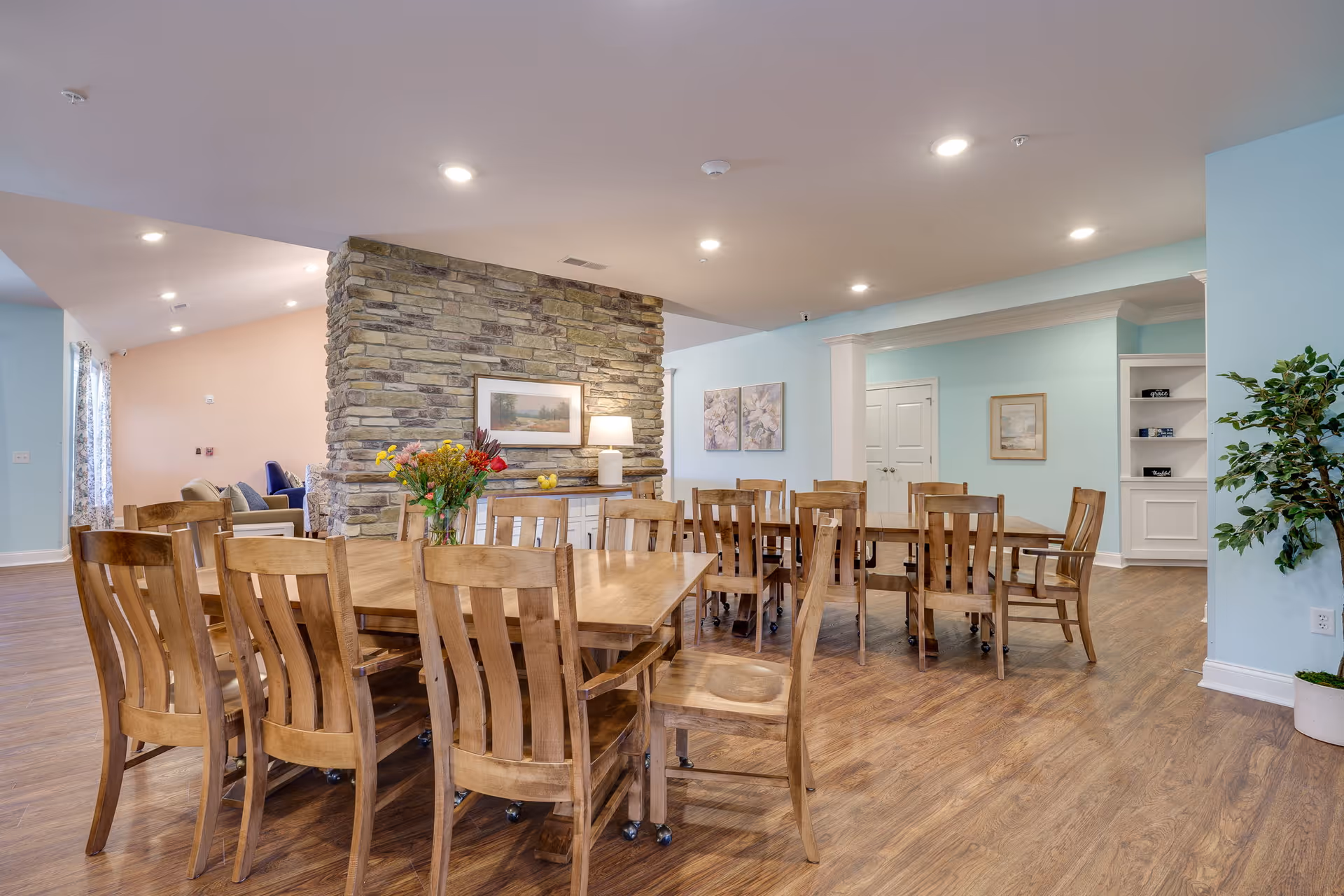 A spacious dining area with multiple wooden tables and chairs on a wood floor. A stone accent wall with a framed picture and a lamp is visible in the background. The walls are painted light blue and beige, with decorative artwork and a potted plant near the corner. The room is well-lit with recessed ceiling lights.