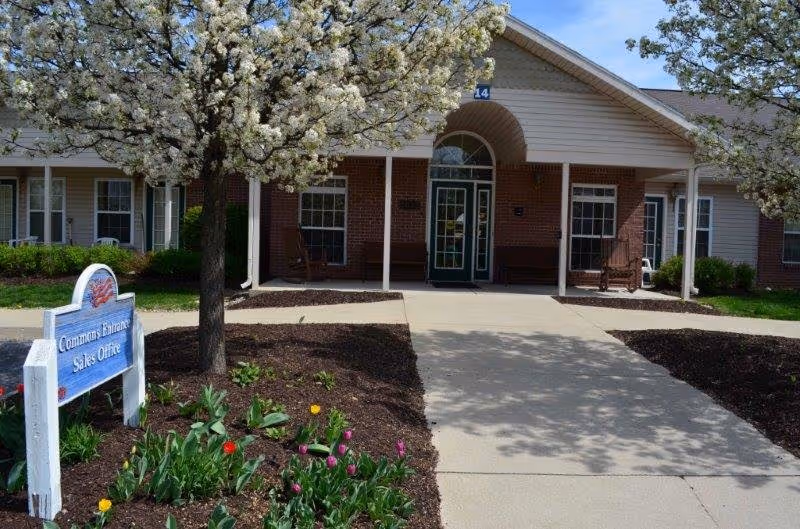 Front entrance of a brick senior living building with a paved walkway, blooming trees, flowerbeds, and a sign reading 'Commons Entrance Sales Office'.