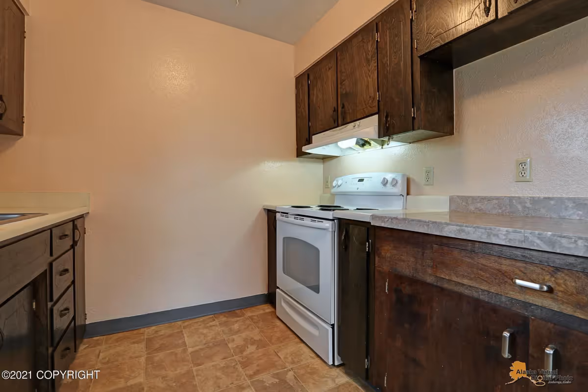 A small kitchen with dark wooden cabinets, a white electric stove with an overhead range hood, beige walls, and a tiled floor. The countertop is light-colored with a marble pattern.