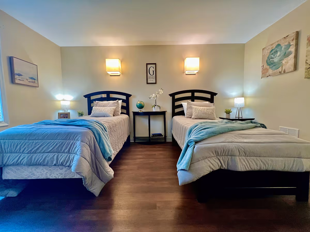 A bedroom with two single beds, each with a light gray comforter and a light blue throw blanket. Between the beds is a small black nightstand with a globe, a white orchid plant, and some books. The walls are beige with two wall-mounted lights above the beds, a framed letter 'G' sign in the center, and decorative artwork on the side walls. There are two table lamps on small side tables next to each bed, and the floor is dark wood.