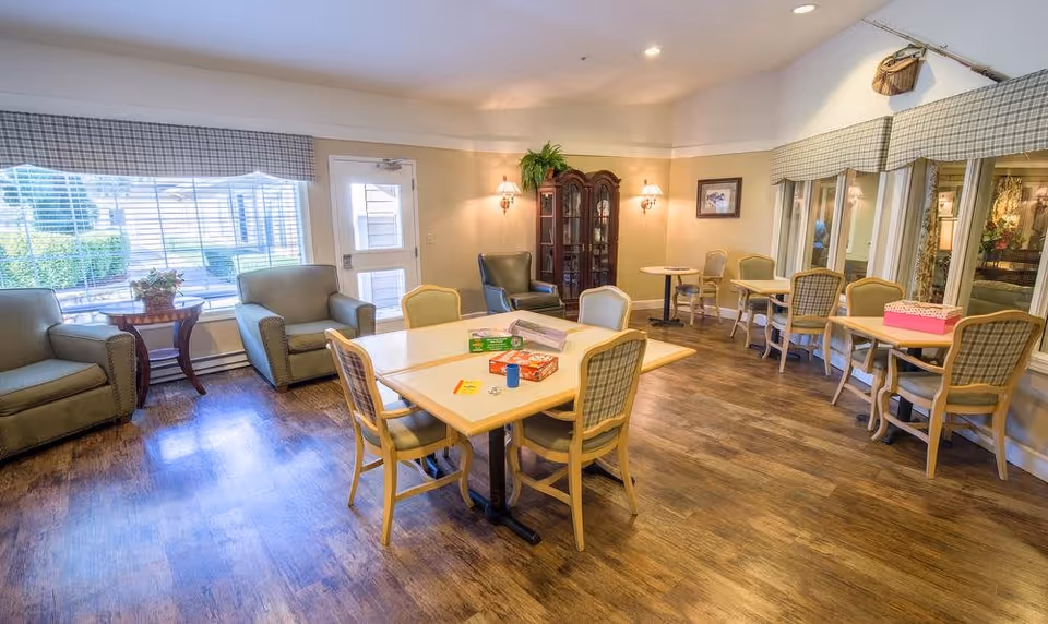 A bright and spacious common area in a senior living facility with multiple tables and chairs arranged for group activities or dining. There are comfortable armchairs near large windows with blinds, a wooden cabinet with glass doors, wall sconces providing warm lighting, and a wooden floor. The room has a cozy and welcoming atmosphere.
