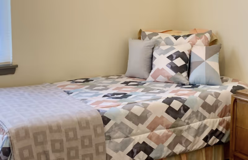 A neatly made bed with a geometric patterned bedspread and several matching pillows in a small bedroom with beige walls and a window with blinds.