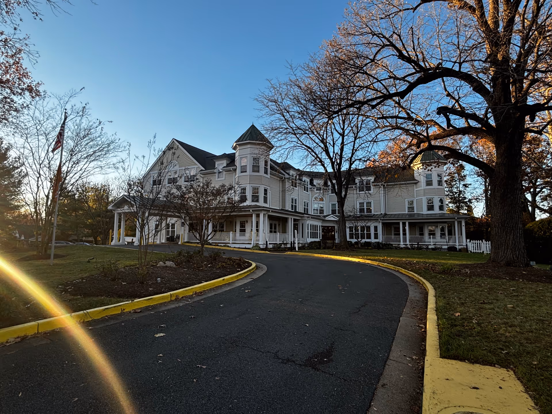 Large three-story Victorian-style building with a wraparound porch, circular driveway and trees under a clear sky.